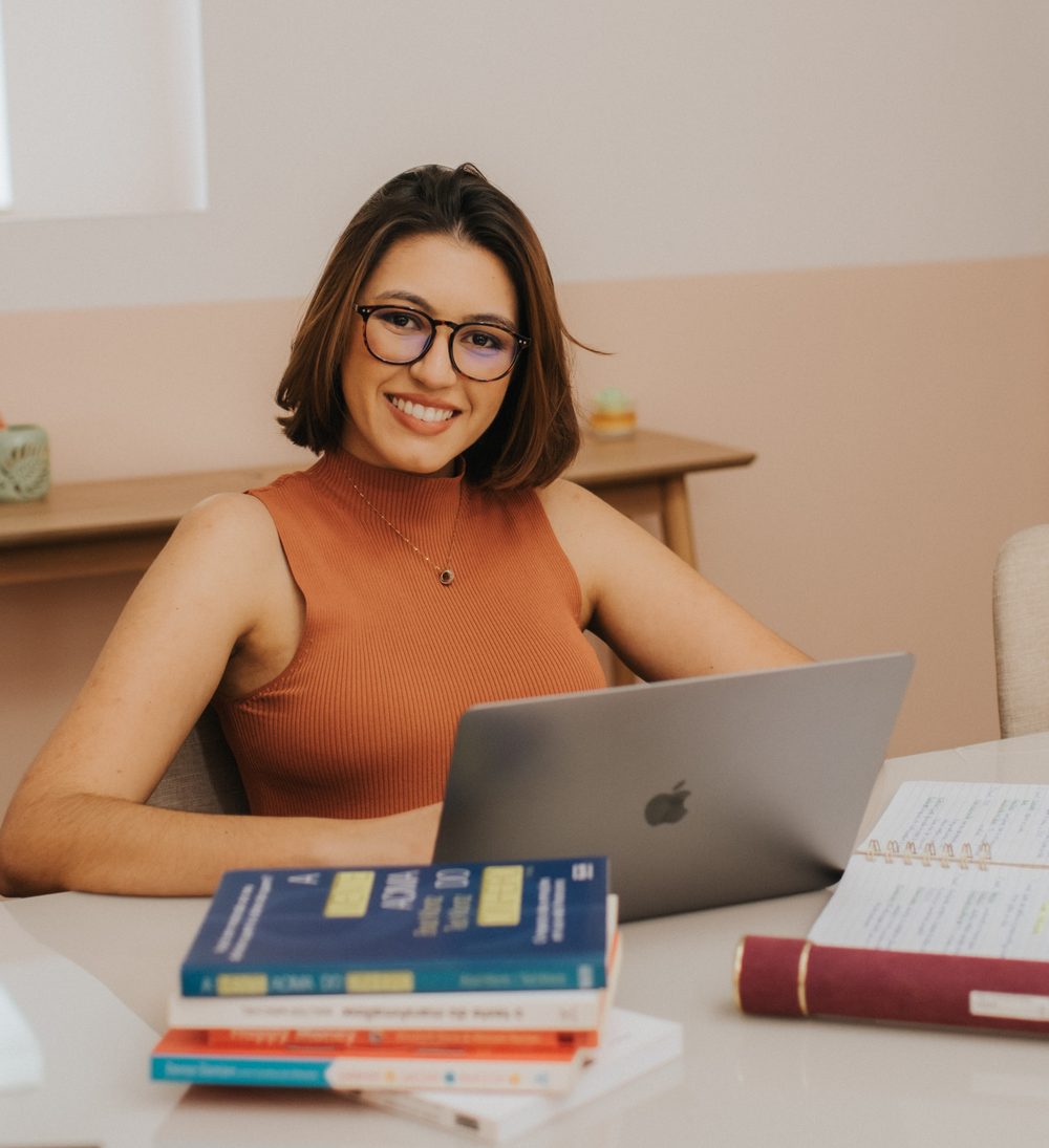 Ana Flávia Vianna sorrindo à mesa de trabalho, com o notebook, livros e o caderno de planejamento